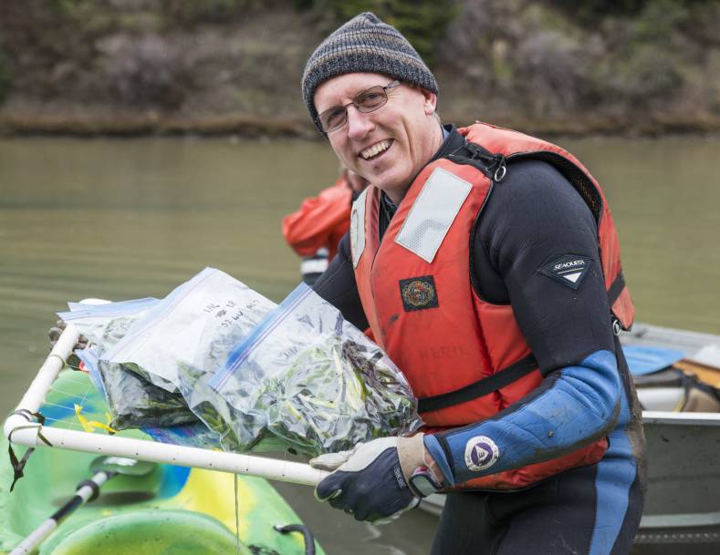 Photo: Dr. Frank Shaughnessy stands next to a boat by the water wearing a wetsuit, life jacket, knit cap, gloves, and glasses. He is holding plastic bags with seaweed.